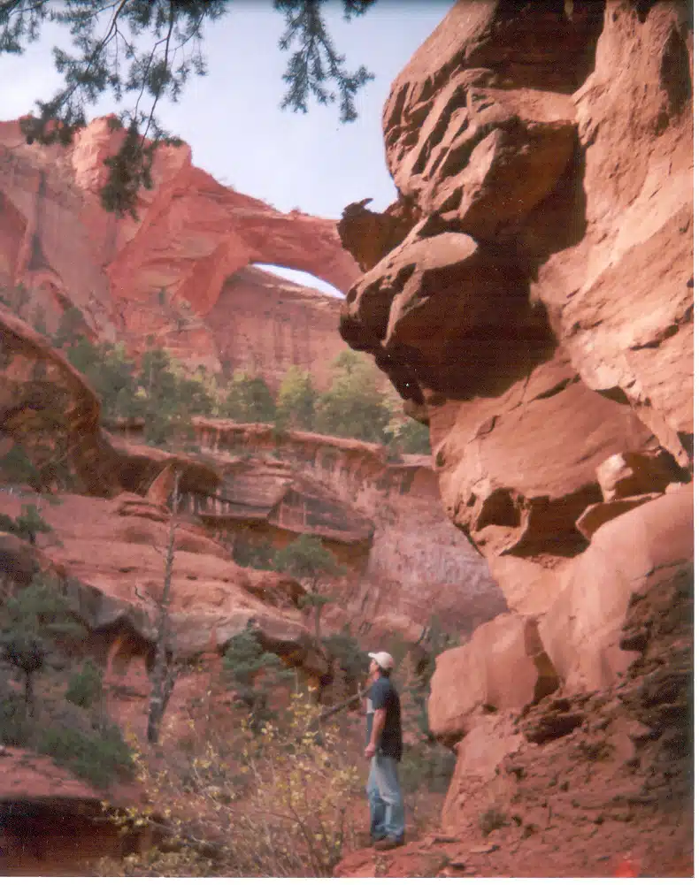 A person stands in a rocky canyon surrounded by red cliffs and greenery.
