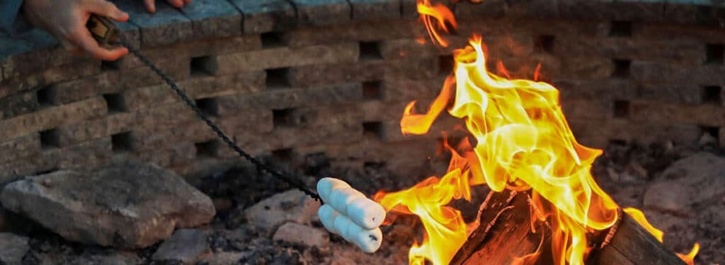 A hand holds a skewer with marshmallows over a crackling fire in a stone pit.