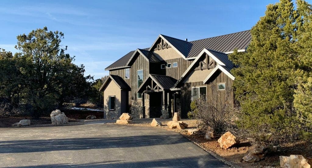 A modern, dark wooden house with a steep roof situated on a gravel driveway surrounded by trees and rocks.