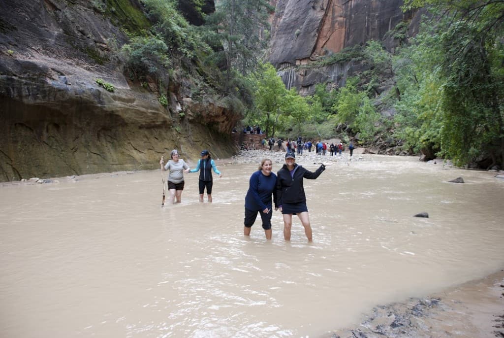 Two women wade through a muddy river in a canyon, smiling at the camera while other hikers are visible in the background.
