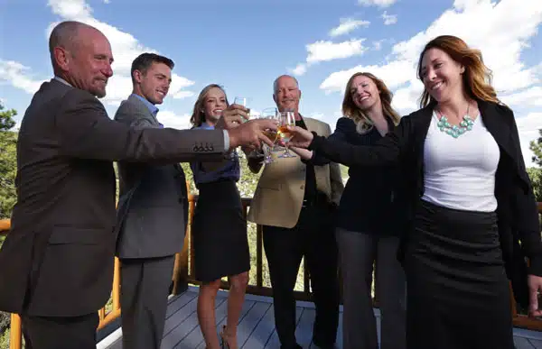 A group of six people in business attire toasting with glasses outside against a scenic background.