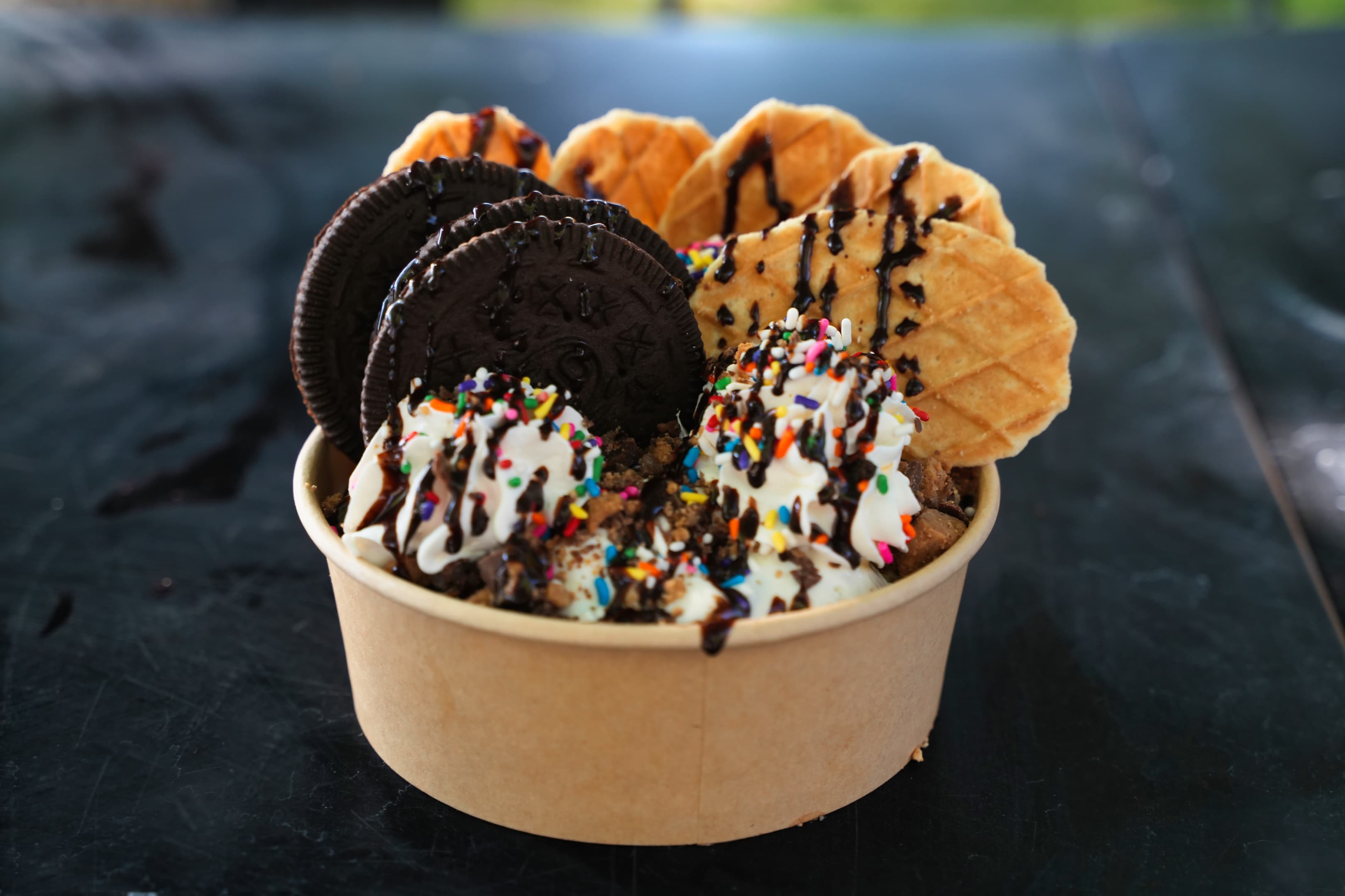 A cup of ice cream topped with whipped cream, sprinkles, chocolate drizzle, Oreo cookies, and waffle cones.