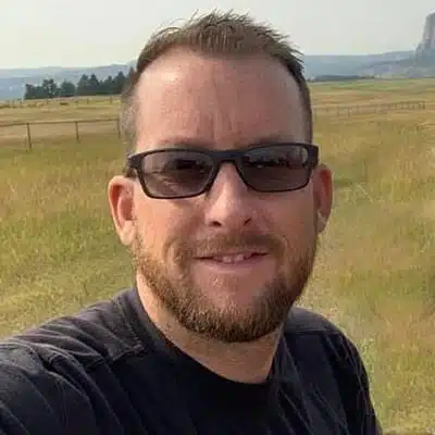 A man with sunglasses smiles in a grassy field with distant hills.