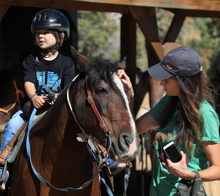 A child in a helmet sits on a horse while an adult woman gently strokes the horse's face.