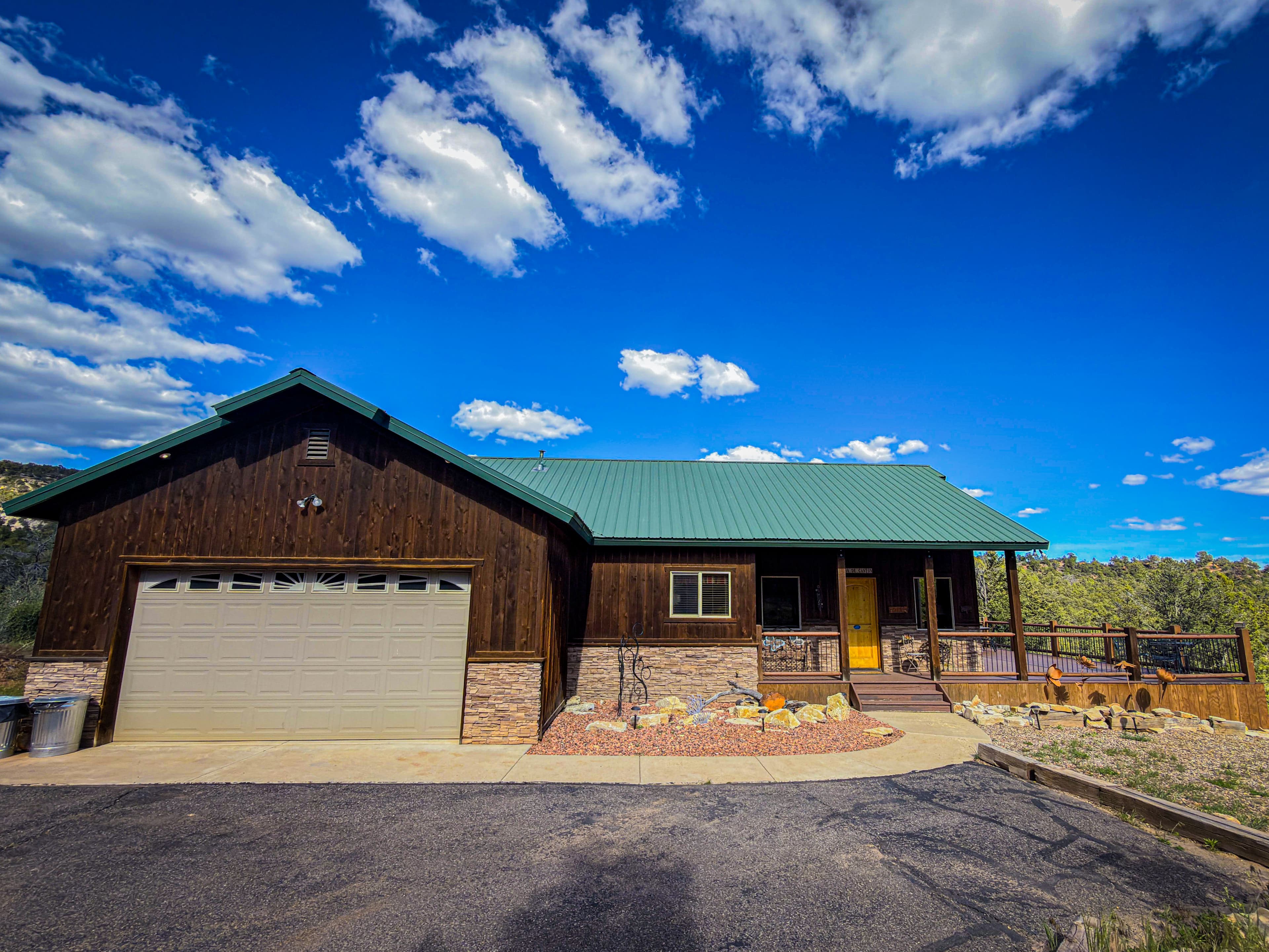 A brown wooden house with a green metal roof and a garage, set against a blue sky with clouds.