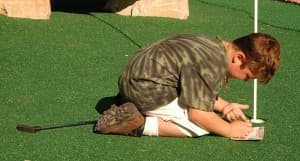 A child kneels on the green grass, focused on writing in a notebook.