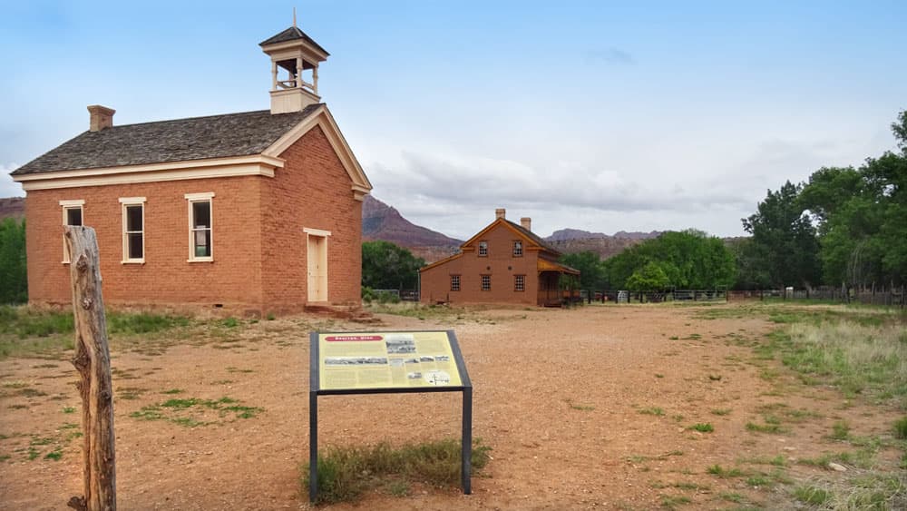 A historic red brick schoolhouse stands next to a larger building, with a sign in the foreground on a gravel pathway.