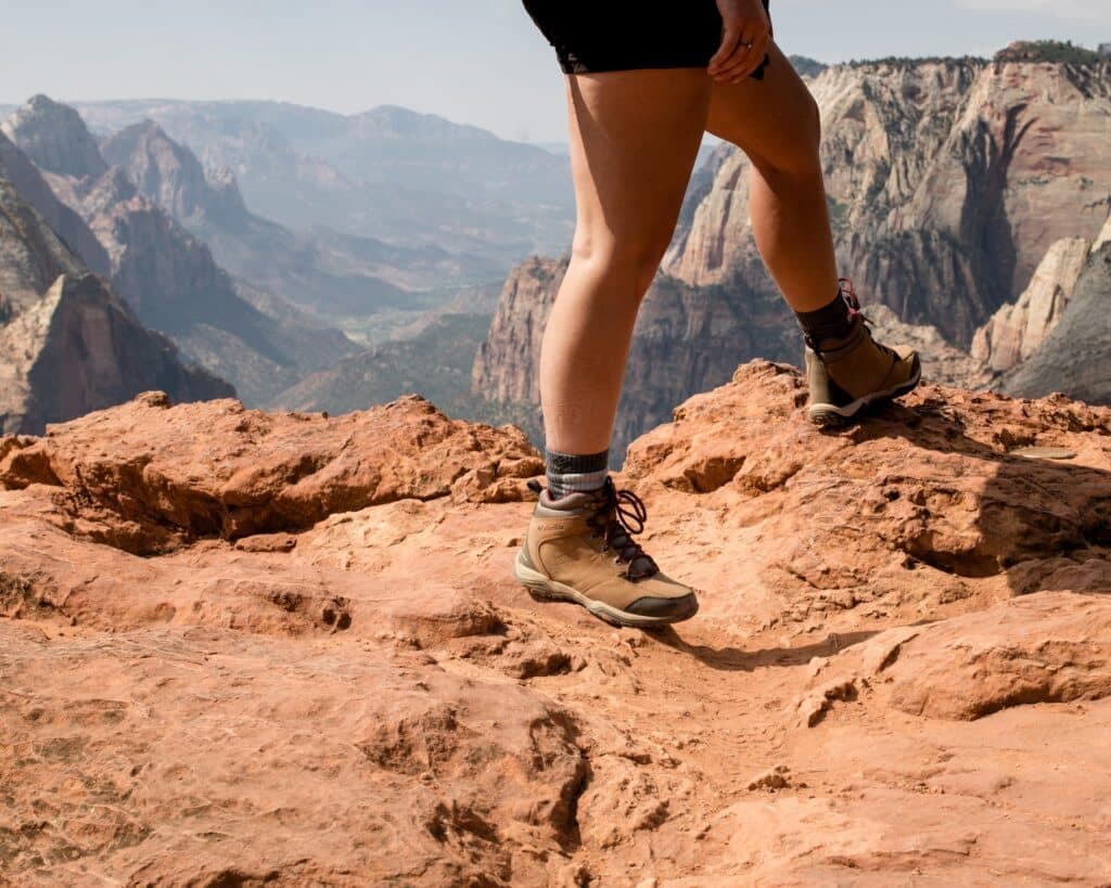 A hiker's leg in sturdy boots stands on a rocky outcrop overlooking a mountainous landscape.
