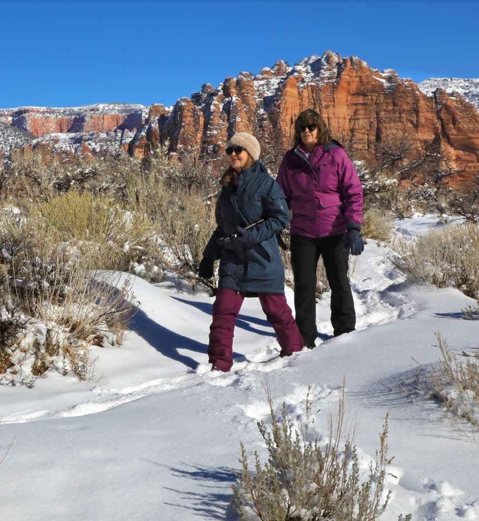 Two women hiking through snow-covered terrain with red rock formations in the background.