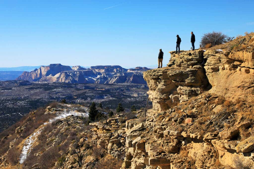 Three people stand on the edge of a rocky cliff overlooking a vast, snowy landscape.