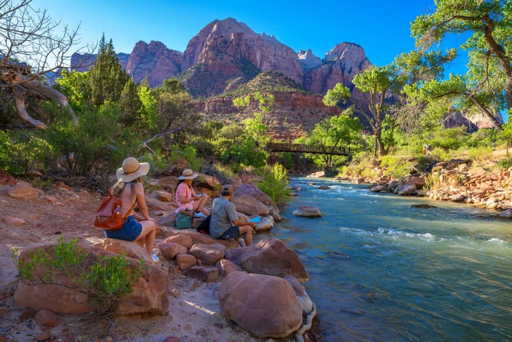 A group of friends sits by a river in a scenic canyon landscape.