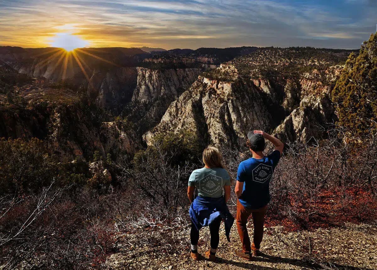 A couple stands on a rocky outcrop, watching the sunset over a canyon landscape.