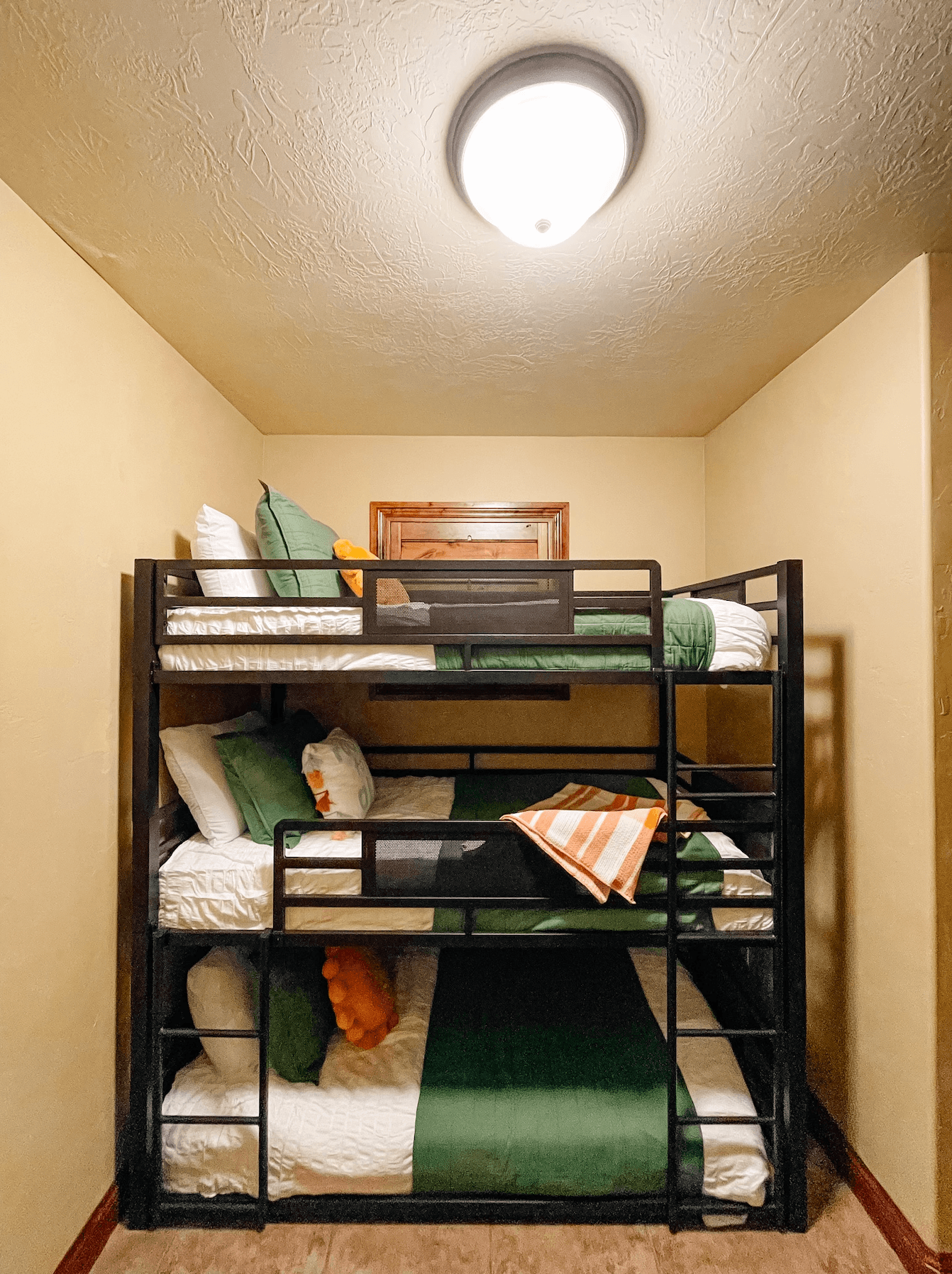 A black metal bunk bed with green and white bedding is set against a beige wall, illuminated by a ceiling light.