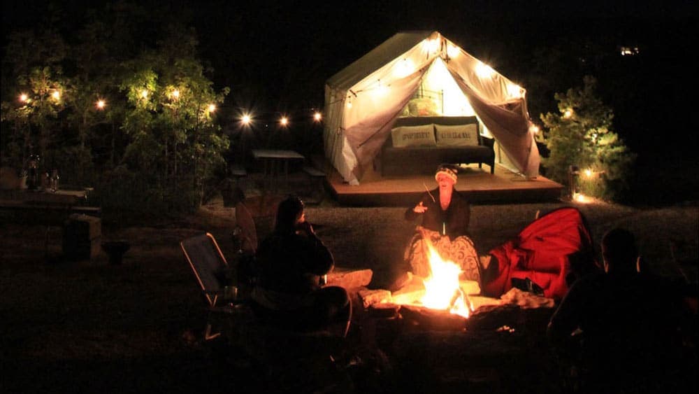 A campfire scene at night with people sitting around and a lit tent in the background.