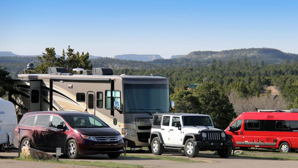 An RV and several parked vehicles are surrounded by a scenic landscape of hills and trees.
