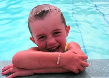 A smiling child leans over the edge of a pool, with her arms crossed and water in the background.
