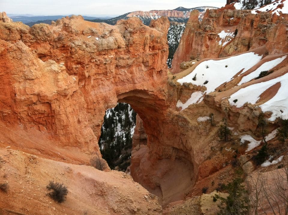A natural arch formed in striking red rock surrounded by snow-covered terrain.