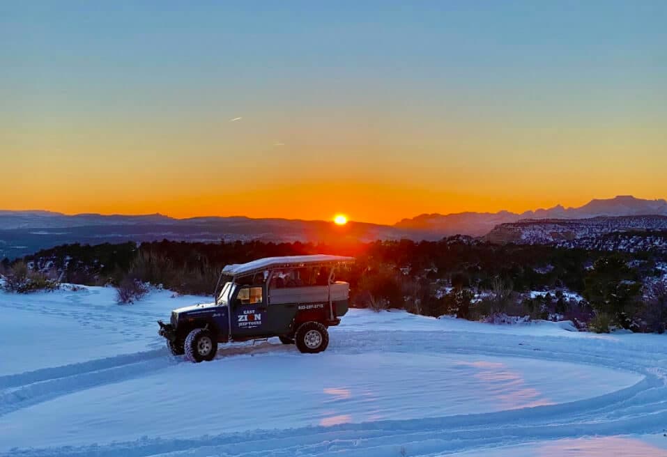 A vehicle parked in snowy terrain at sunset, with mountains in the background.