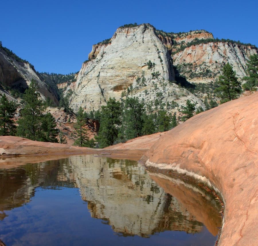 Reflective pool in front of towering sandstone cliffs under a clear blue sky.