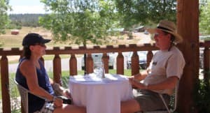 A woman and a man are sitting at a table on a sunny patio, engaging in conversation amid green trees.
