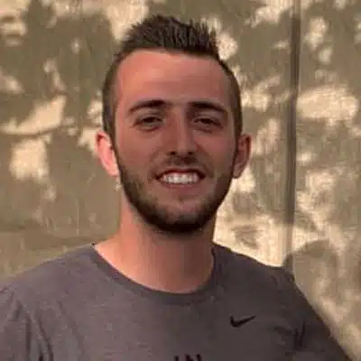 A young man with short hair and a beard smiles while standing outside.