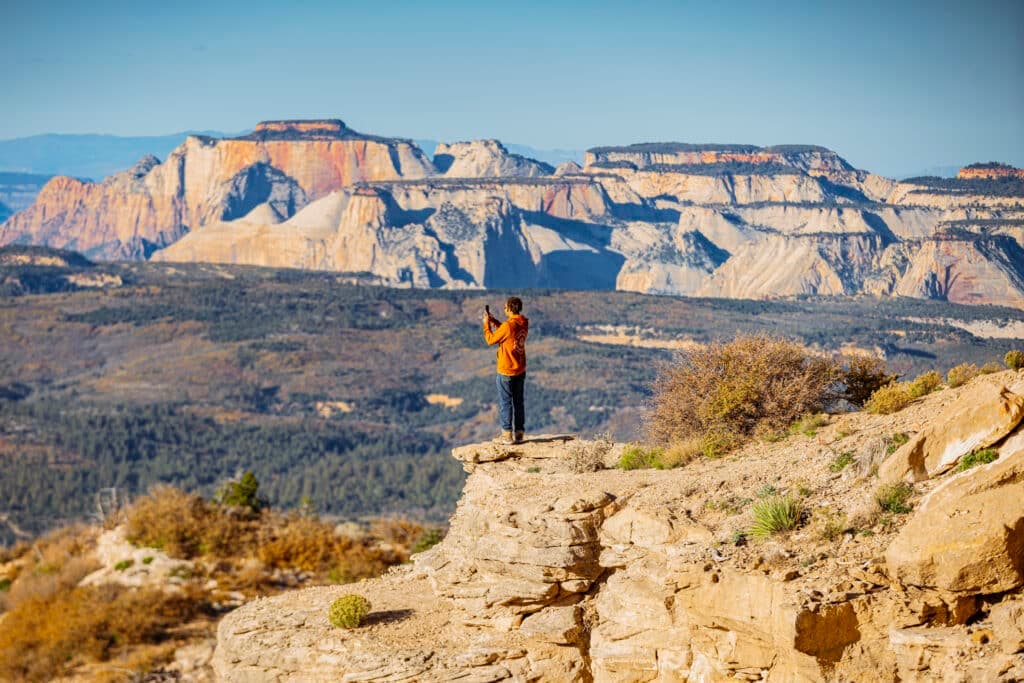 A person in an orange jacket stands on a rocky ledge, taking a photo of expansive mountains in the background.