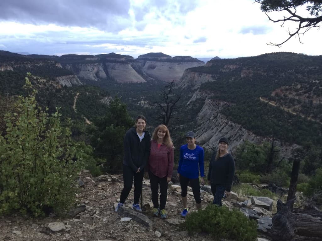 Four people pose for a photo on a mountainous landscape with lush greenery and a cloudy sky.
