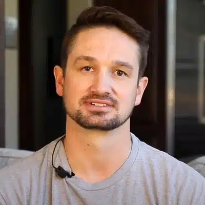 A man with short hair and a beard, wearing a gray shirt, sits in a well-lit room.