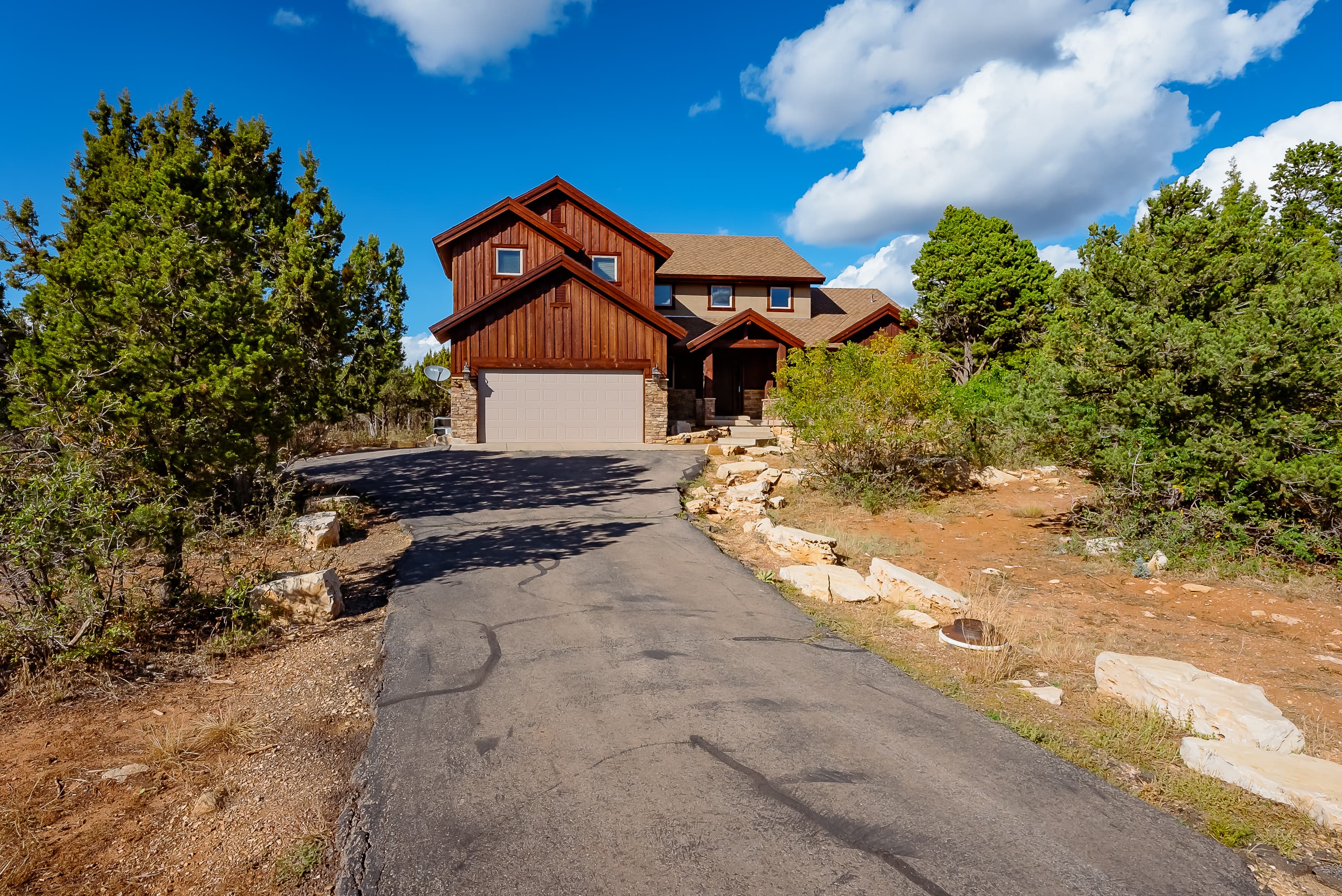 A large wooden house with a sloped roof and a paved driveway surrounded by greenery and boulders.