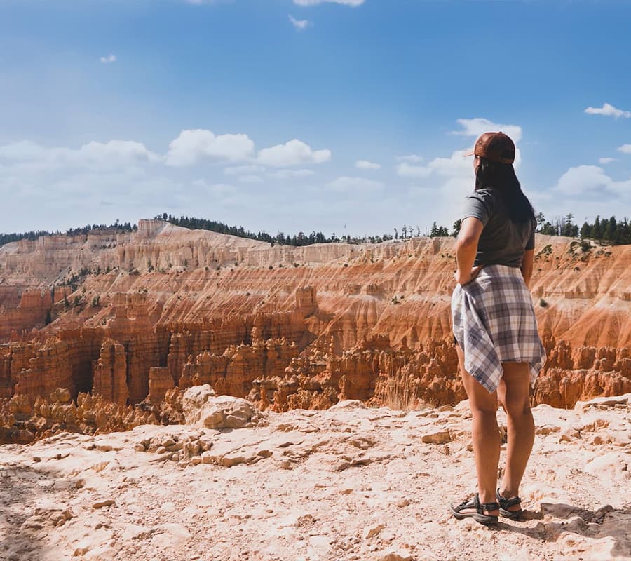 A person stands on a rocky ledge, gazing at the expansive, orange-hued canyon under a blue sky.