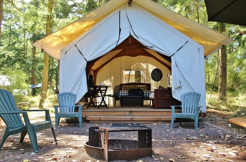 A cozy tent setup with chairs and a fire pit, surrounded by trees.