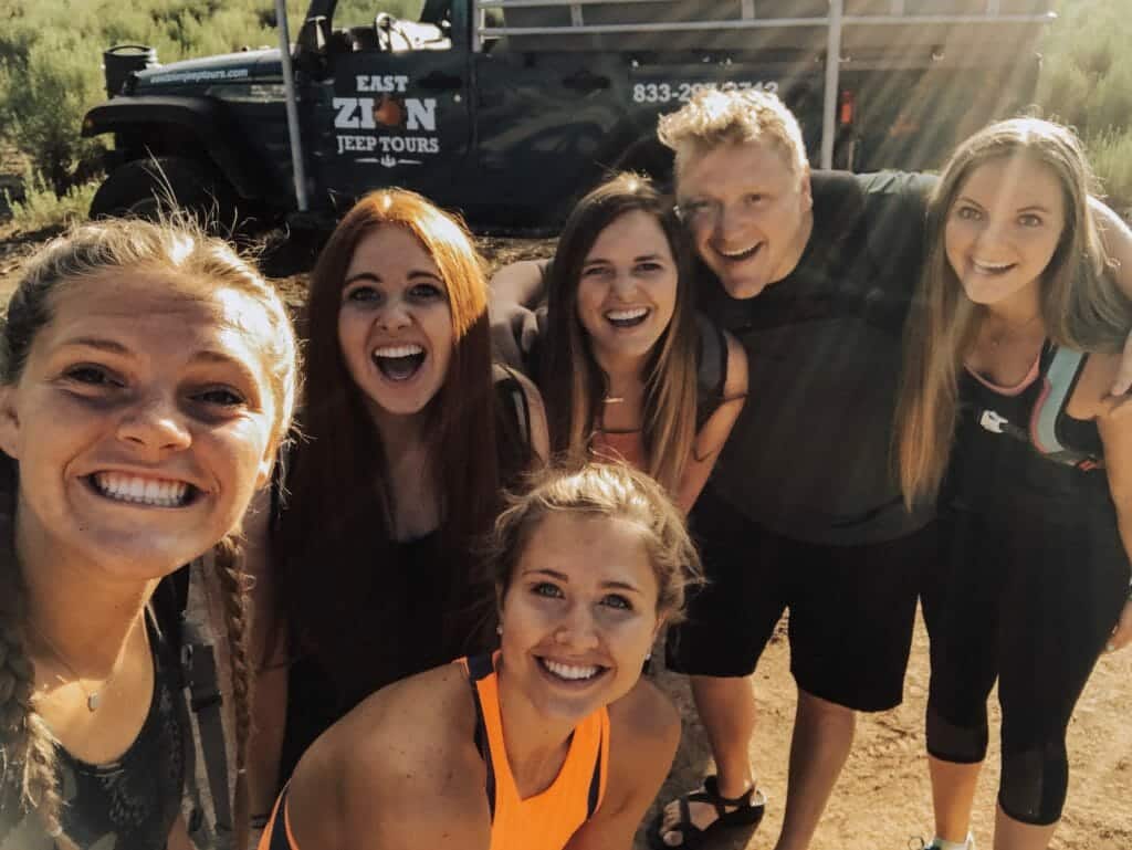 A group of six friends smiles for a selfie with a jeep in the background.