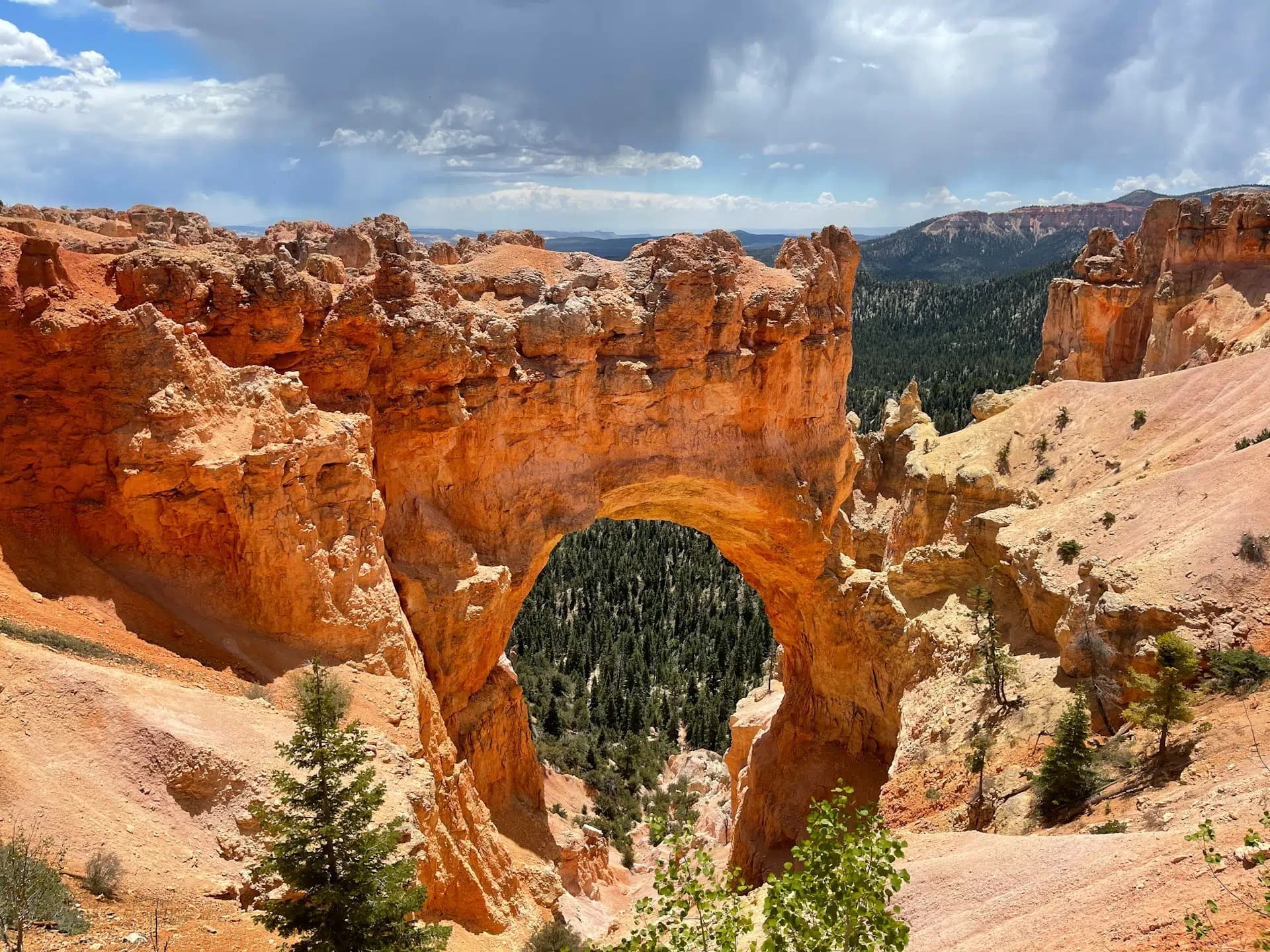 A natural rock arch surrounded by rugged terrain and a forested valley under a cloudy sky.