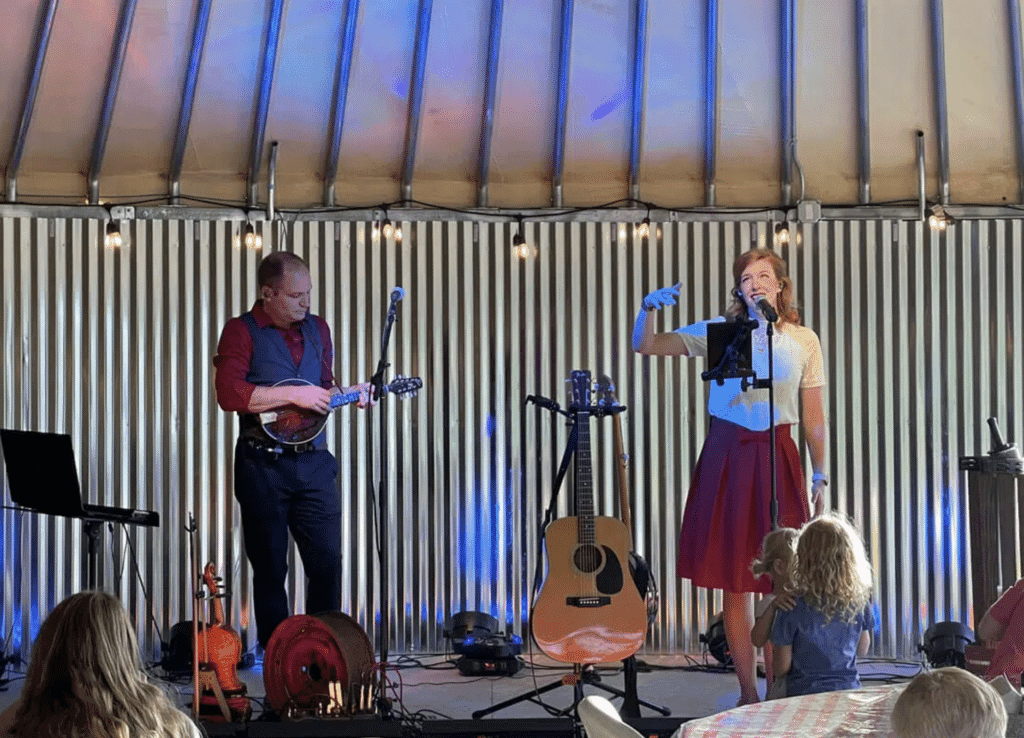 A man plays a mandolin while a woman sings on stage in a cozy venue.