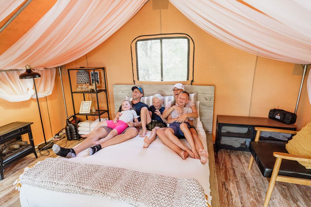 A group of five children and adults sit closely on a bed inside a cozy tent.