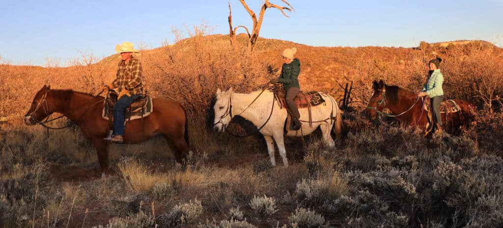 Three riders on horseback traverse a desert landscape at sunset.