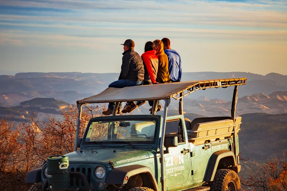 Four people sit on the roof of a jeep, enjoying a scenic view of mountains at sunset.