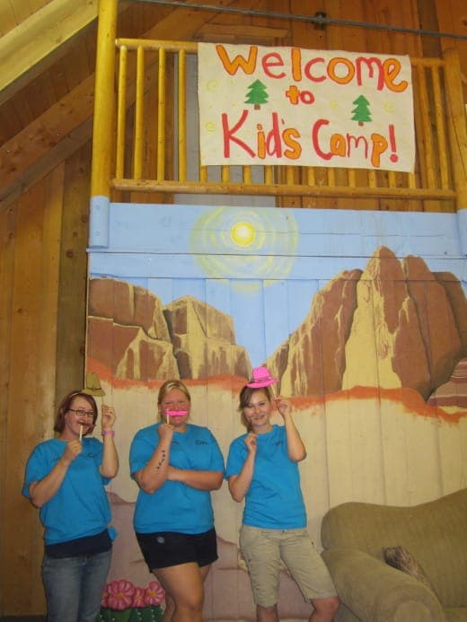 Three camp staff members in blue shirts pose playfully in front of a "Welcome to Kids Camp!" banner and a colorful mural.
