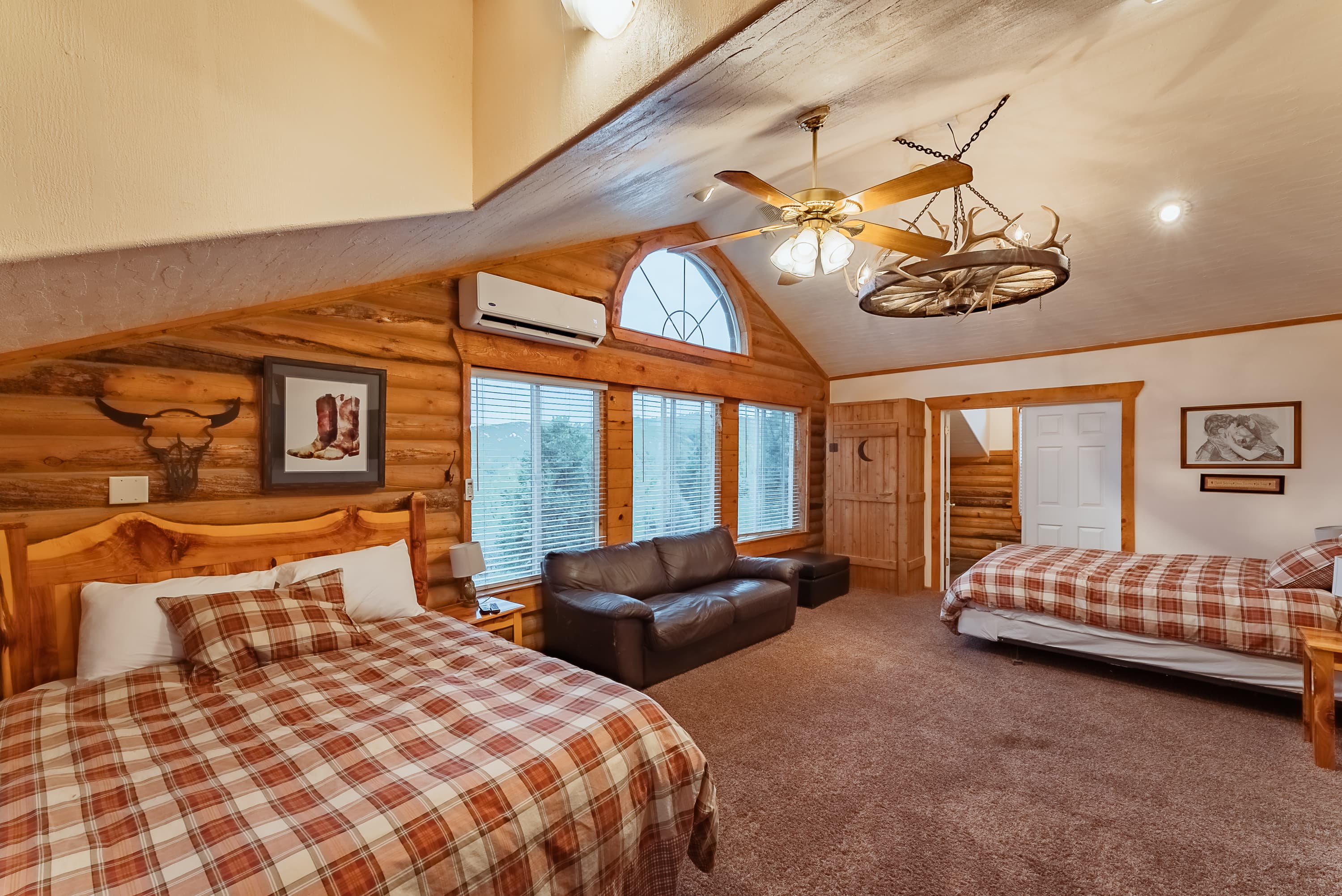 A cozy rustic bedroom featuring two double beds, a leather sofa, and large windows with wooden accents.