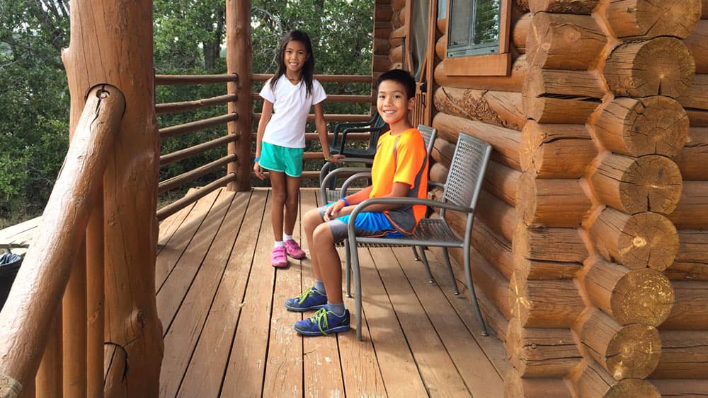 Two children, a girl in a white shirt and shorts and a boy in an orange shirt and blue shorts, smile while standing and sitting on a wooden porch.