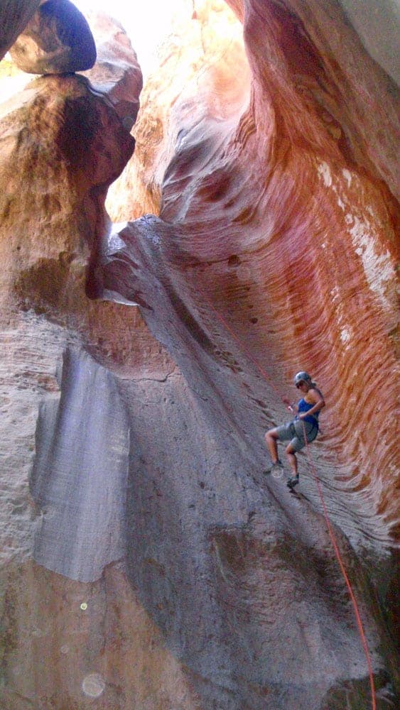 A person rappels down a steep, narrow canyon with colorful rock formations.