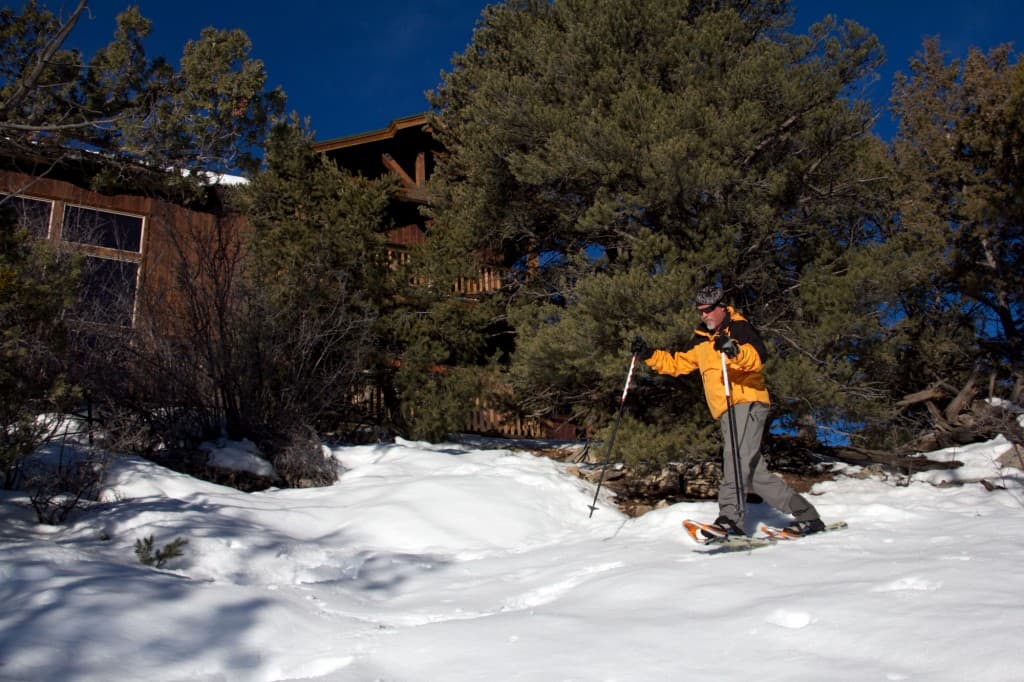 A person in an orange jacket snowshoes through snowy terrain near a wooden cabin.