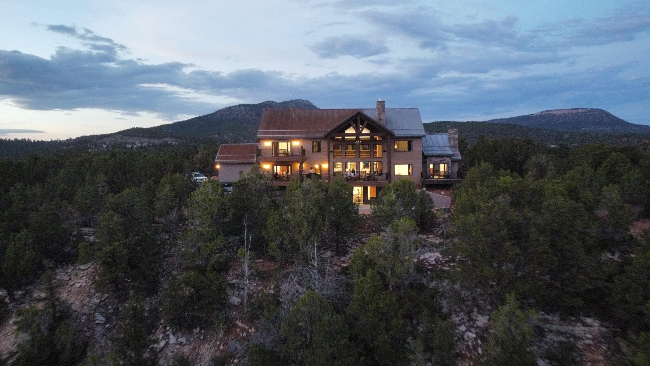 A large, rustic house sits among trees, with mountains visible in the background under a cloudy sky.