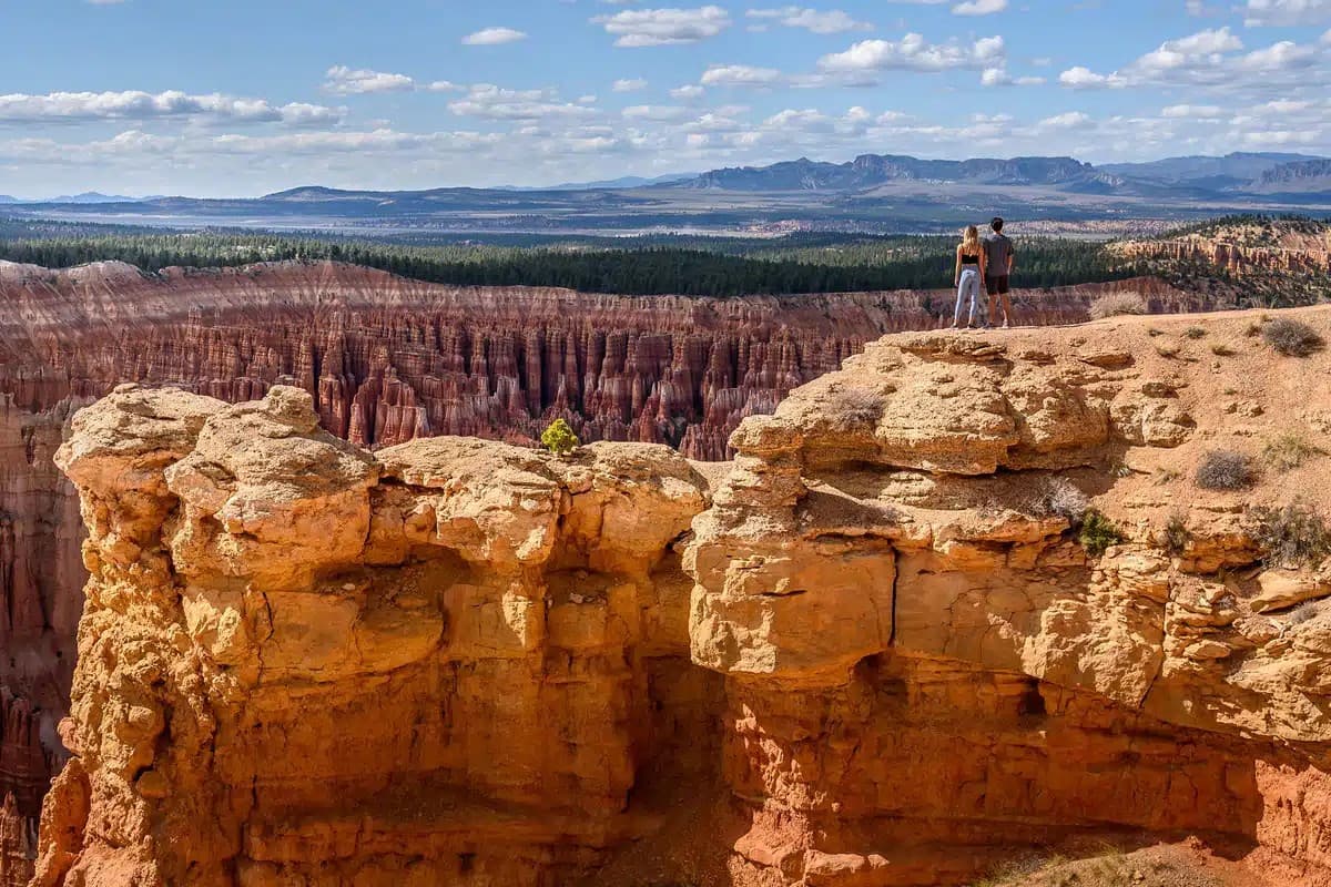 A couple stands on a rocky outcrop overlooking a vast canyon landscape under a blue sky.