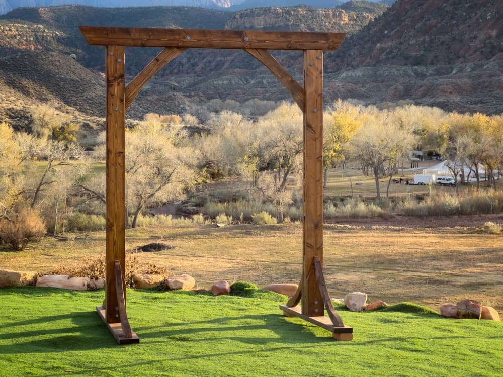 A wooden arch stands on green grass with a scenic landscape and mountains in the background.