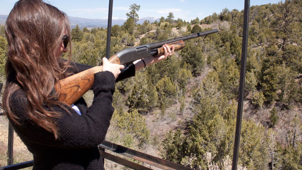 A person aiming a shotgun at a distant landscape from a shooting platform.