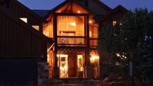 A warmly lit wooden house at dusk, featuring a front porch and large windows.