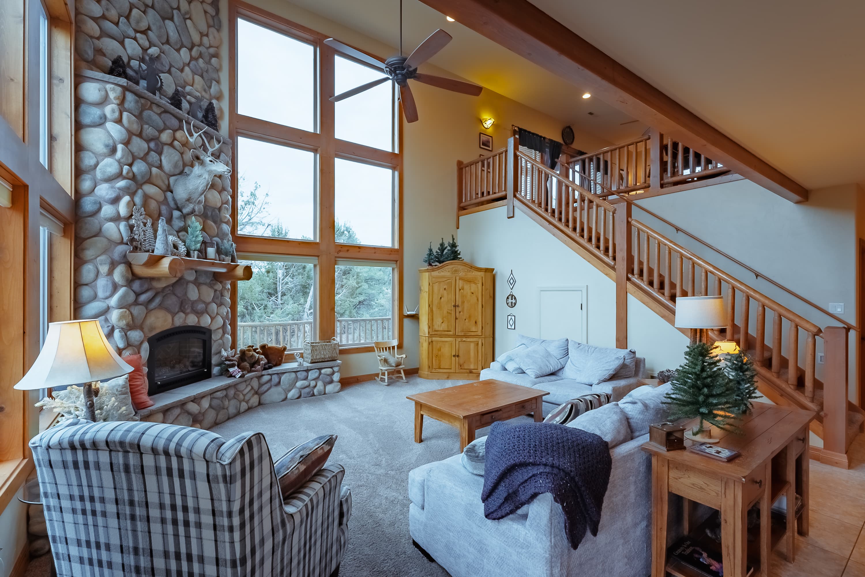 A cozy living room with large windows, a stone fireplace, and a wooden staircase.