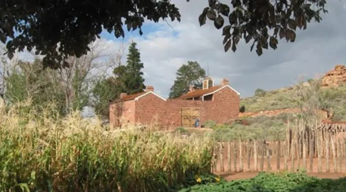 A rustic red brick building surrounded by lush greenery and cornfields under a cloudy sky.