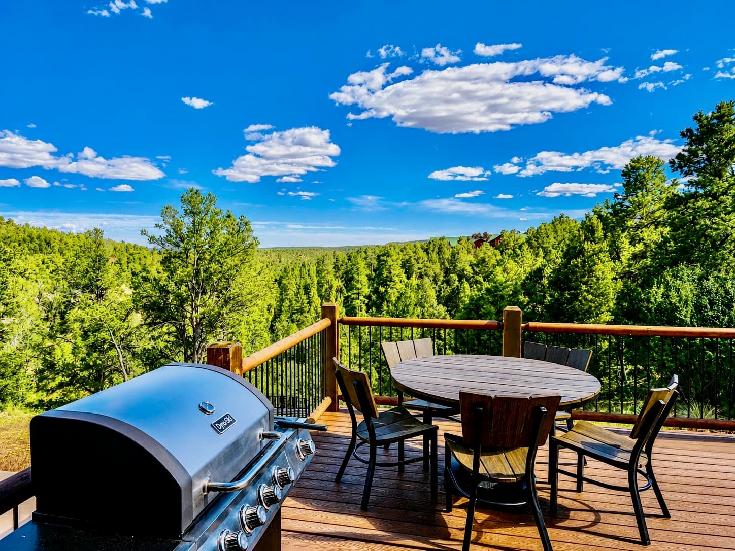 A deck with a grill and outdoor seating overlooks a lush green landscape under a blue sky with clouds.